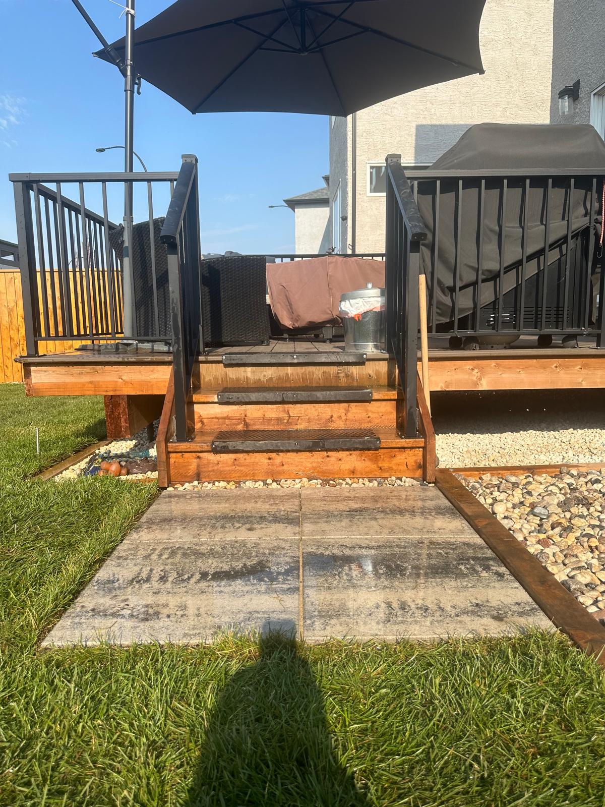 A wooden deck with black metal railings and steps featuring anti-slip treads leads to a patio surrounded by decorative pebbles and grass. An umbrella shades outdoor furniture on the deck, with stone slabs forming a path from the lawn to the steps