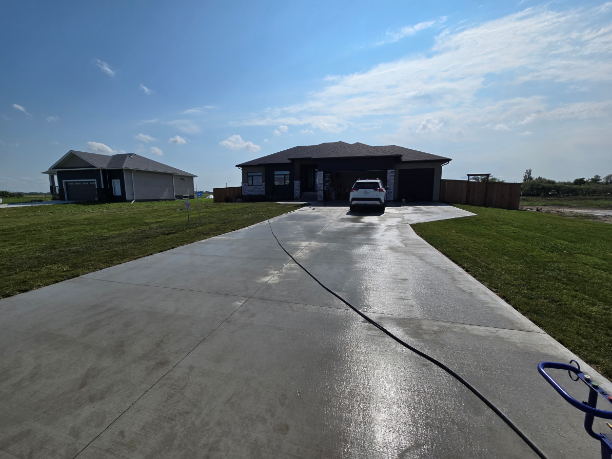 A long concrete driveway, bordered by fresh grass, leads to a modern, single-story house with a parked white car near the garage. A hose lies on the driveway under bright, sunny skies, suggesting recent cleaning.